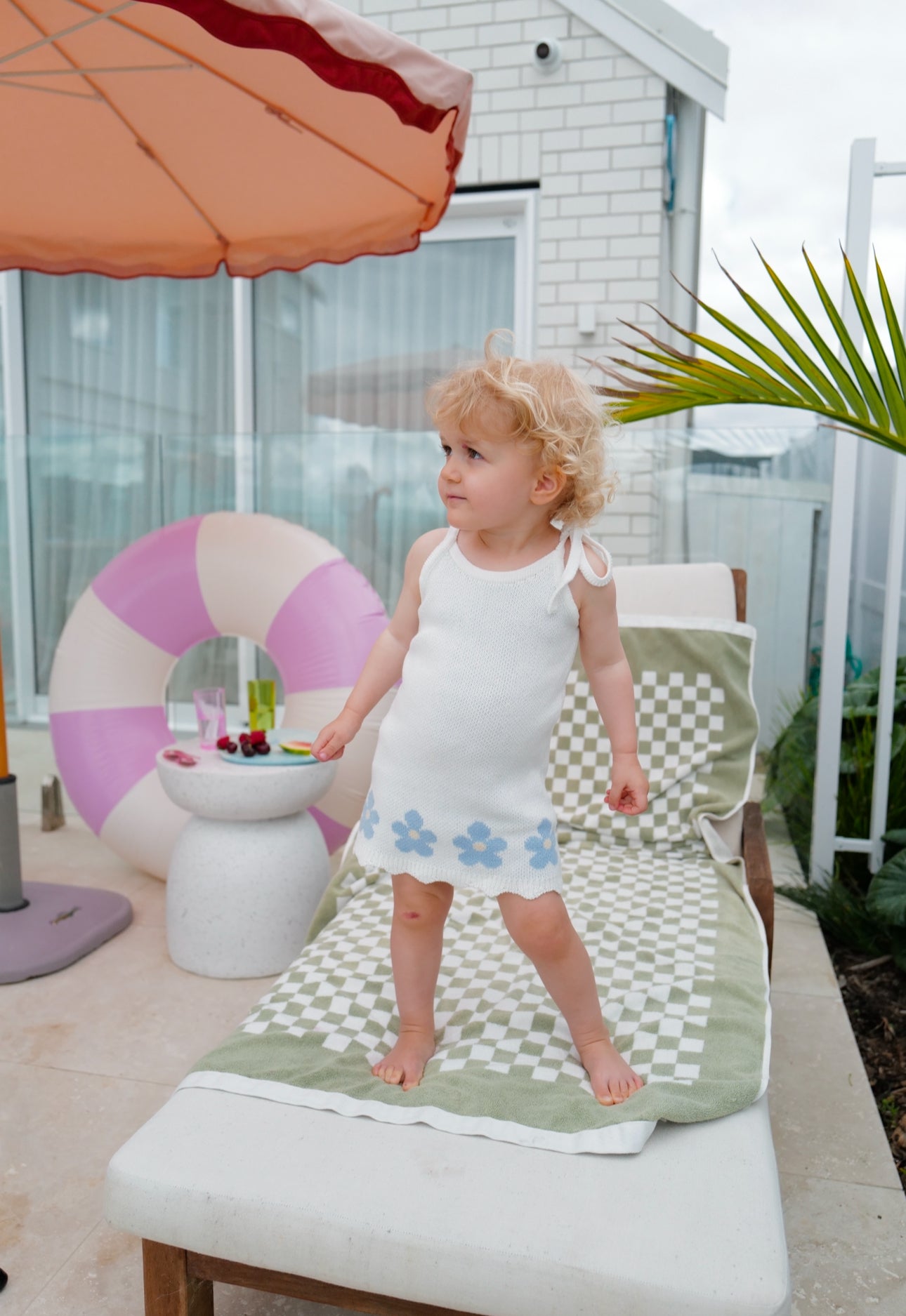 Toddler stands on lounger with umbrella and pool toys in background. Wearing blossom knit dress