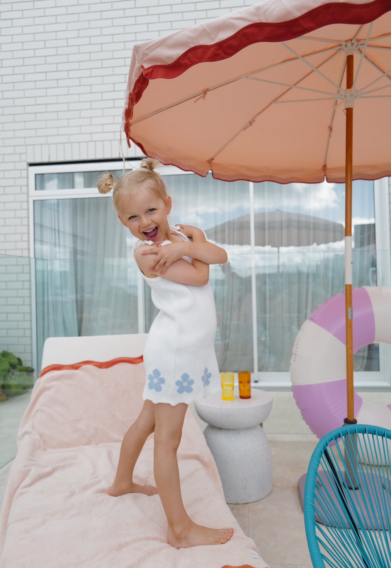 Girl stands on lounger with umbrella and pool toys in background, happy expression holding arms across her body. Wearing blossom knit dress